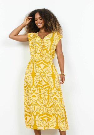 Frau mit lockigem Haar, die ein ärmelloses Midikleid mit gelbem und weißem Muster trägt und lächelt, während sie mit der Hand ihr Haar berührt.