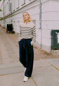 Striktrøje med cremefarvede og sorte striber, halv-zippelukning; navy kord- bukser med bred ben; hvide sneakers; urbant miljø med brostensbelagt sti.