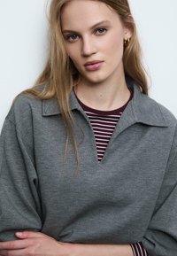 Grå kravepullover med V-hals, lagd over en maroon og hvid stribet skjorte. Blød tekstur, semi-løs pasform og afslappet design.