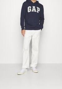 Man i en marinblå GAP-huvtröja, vita byxor med uppvikta ben och vita sneakers står mot en vit bakgrund.