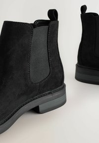 Botas negras de tobillo con un panel lateral elástico texturizado y un tacón bajo, situadas sobre un fondo neutro.