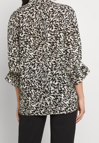 Leichte, durchsichtige Bluse mit einem schwarz-weißen abstrakten Muster, ausgestattet mit einem Kragen und rüschigen Säumen, kombiniert mit dunklen Hosen.