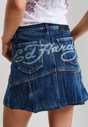 Vue de dos d'une personne portant une jupe en denim bleu avec deux poches et le texte "Ed Hardy" au dos.