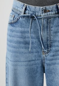 Light blue denim jeans with a drawstring waist, zip fly, and five pockets. Features subtle fading and contrasting stitching details.
