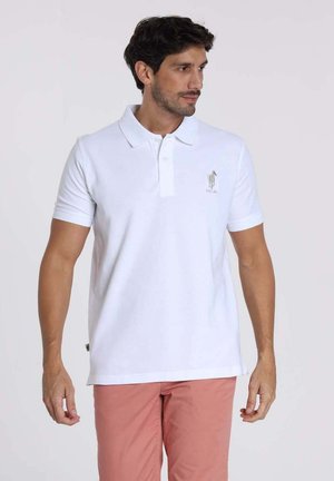 Polo blanc en coton, doté d'un col court, de deux boutons et d'un logo brodé sur la poitrine gauche. Porté avec un pantalon rose clair.