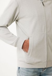 Svetle šedá mikina na zip s texturovanou tkaninou, ribbovanými manžetami a dvěma bočními kapsami. Zip má kovový táhlo.