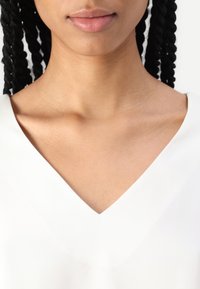 Bílý top s výstřihem do V a hladkou texturou, který prezentuje čistý design a výrazný límec. Blízko střižený, zaměřuje se na oblast krku a horní části hrudi.