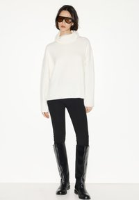 Witte, grofgebreide coltrui, zwarte legging en zwarte, kniehoge laarzen. Model draagt oversized donkere zonnebril en poseert tegen een witte achtergrond.