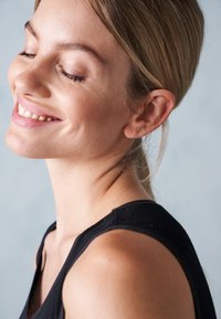 Sort ærmeløs top, glat stof. Lysebrunt hår trukket tilbage. Blød hudtone med minimal makeup. Nærbillede af ansigt og nakke.