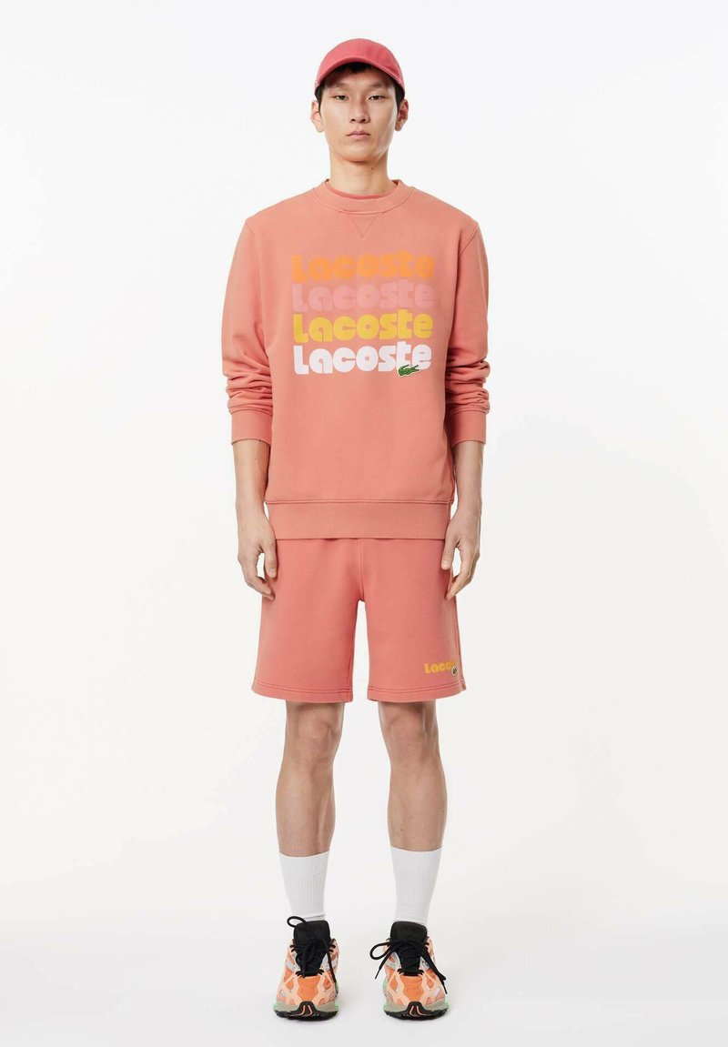 Felpa rosa con logo "Lacoste" in vari colori, pantaloni corti rosa abbinati, calzini bianchi e scarpe da ginnastica multicolori con motivo testurizzato.