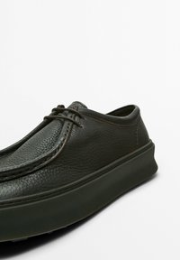Chaussure en cuir vert foncé avec un bout arrondi, une surface texturée, des coutures discrètes et une épaisse semelle en caoutchouc noir. Les lacets ajoutent une touche de détail.