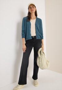 Cardigan teal, sous-vêtement léger, jeans noirs taille haute, baskets beiges et un sac crème. Texture lisse, accents de boutons sur le cardigan.