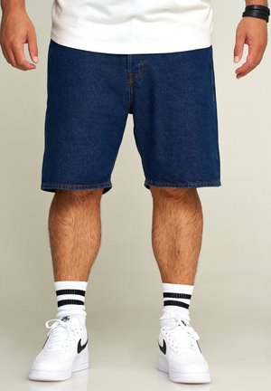 Denim shorts in dark blue with a straight cut, raw edges at the hem, and contrast stitching. Paired with white sneakers and black-striped socks.
