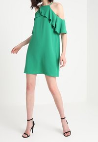 Abito verde con spalle scoperte e dettagli in volant, realizzato in un tessuto leggero. Presenta una silhouette ampia e un orlo sopra il ginocchio.