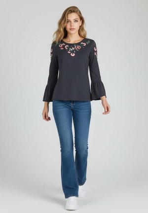 Mujer caminando con una blusa negra con bordado floral y mangas campana, jeans acampanados azules y zapatillas blancas sobre un fondo liso.