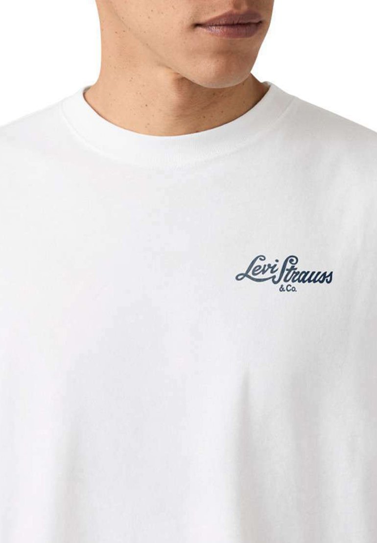 Hombre con camiseta blanca de cuello redondo con pequeño logo azul oscuro "Levi Strauss & Co." en el pecho izquierdo, labios y línea de la mandíbula visibles.