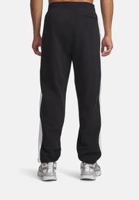 Mężczyzna w czarnych spodniach dresowych z białymi panelami po bokach i srebrnych butach sportowych, pokazany od tyłu na jednolitym tle.