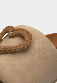 Κοντινό πλάνο μπεζ παπουτσιού από σουέντ με πλεκτή καρδιά από δέρμα σε σκούρο ταμπά χρώμα και μικρό χρυσό κούμπωμα στην κορυφή.