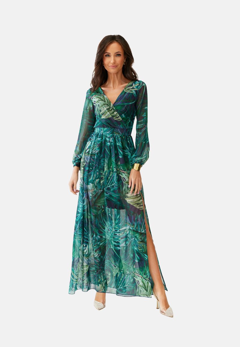 Žena v dlouhých šatech se zeleným potiskem tropického listí, s dlouhými průhlednými rukávy a bočním rozparkem, v kombinaci se zlatými podpatky a zlatým náramkem na zápěstí.