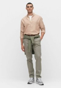 Lichtbruine linnen overhemd met opgerolde mouwen, olijfgroene chinos en een groene gebreide trui om de taille geknoopt, gecombineerd met grijze sneakers.