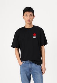 Camiseta negra de manga corta con un gráfico de círculo rojo en el pecho. Fabricada en algodón ligero, presenta un corte relajado. Llevada con jeans azules.