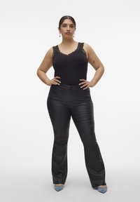 Zwarte bodysuit met kant, gecombineerd met glanzende zwarte bootcut broek. Model draagt blauwe hakken, met de armen gekruist, voor een neutrale achtergrond.