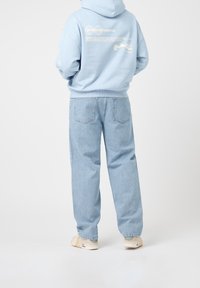 Felpa con cappuccio azzurro chiaro con testo e grafiche stampate, abbinata a jeans azzurro chiaro a vestibilità morbida e sneakers beige su uno sfondo neutro.