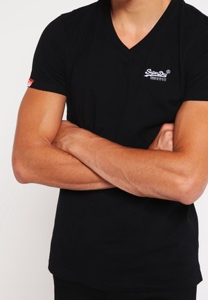 Homme portant un T-shirt noir à col en V, les bras croisés, affichant le logo « Super Dry » sur la poitrine et une petite étiquette rouge sur la manche.
