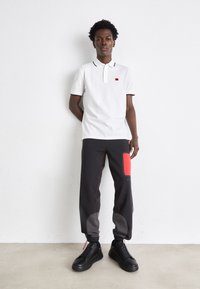 Polo bianco con rifiniture nere e logo rosso, abbinato a pantaloni jogger grigio carbone con accenti rossi e texture variegate, completato da sneakers nere.
