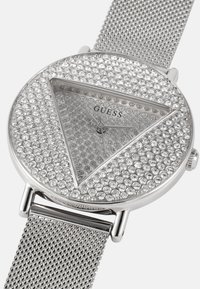 Reloj con correa de malla plateada que presenta una caja redonda adornada con cristales transparentes, esfera triangular y un dial minimalista con agujas plateadas.