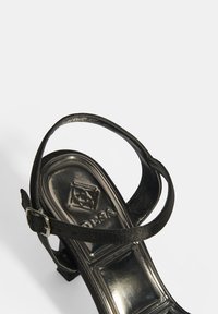 Schwarze High Heels Sandalen mit breitem Riemen, verstellbarem Schnallenverschluss und glatter, glänzender Sohle mit geprägt Logo-Detail.