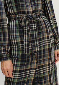 Detailný záber na kárované šaty s dlhými rukávmi, siahajúcou prednou časťou so zapínaním na gombíky a viazaným textilným opaskom v oblasti pása, s rukami položenými po boku.