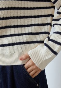 Pull rayé crème et bleu marine en maille avec poignets et ourlet côtelés, tissu texturé, porté sur un denim foncé, main dans la poche.