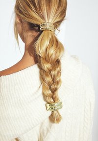 Fermagli dorate che tengono alta una coda di cavallo intrecciata; texture liscia, design a strati con una leggera lucentezza. Maglione soffice color avorio sullo sfondo.