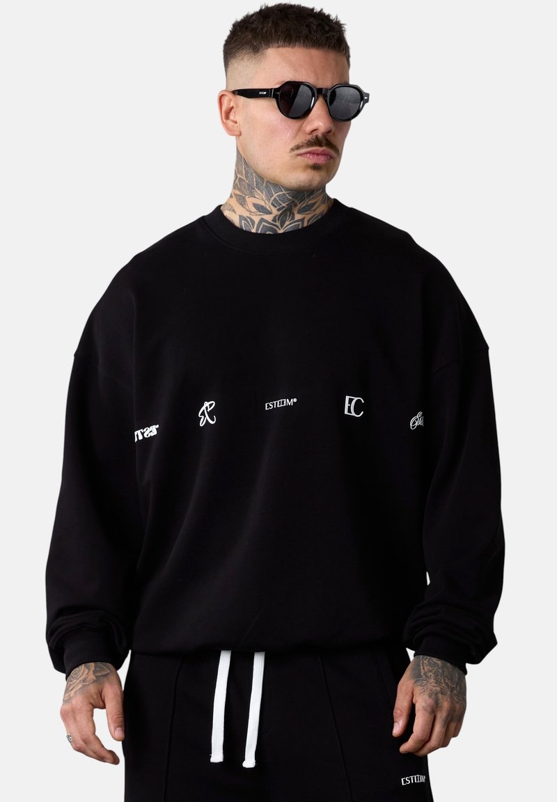 Sweatshirt noir surdimensionné avec des logos blancs imprimés à l'avant. Le tissu semble doux avec des poignets côtelés. Associé à un pantalon noir et un cordon de serrage blanc.