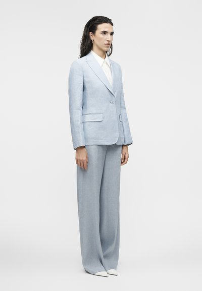 Γυναίκα με μακριά σκούρα μαλλιά, φορώντας ανοιχτό μπλε κοστούμι προσαρμοσμένης γραμμής με παντελόνια φαρδιάς γραμμής και λευκά μυτερά παπούτσια, στέκεται μπροστά σε έναν απλό φόντο.