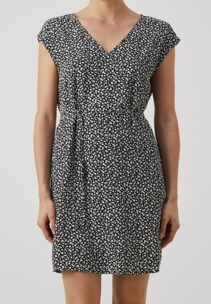 Woman wearing a short black dress with small white floral patterns and a V-neck, standing against a plain light background.