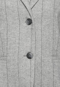 Manteau en laine gris à chevrons avec boutons noirs, col cranté et détails de coutures verticales sur le devant.