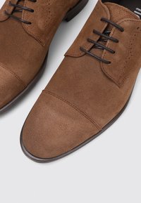 Braune Wildleder-Schnürschuhe mit einer glatten Textur, runder Spitze und kontrastierenden dunklen Lederakzenten an den Ösen und der Sohle.