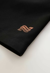 Černý textilní top s hladkou texturou, s měděným logem v dolním rohu a čistě prošitými okraji.