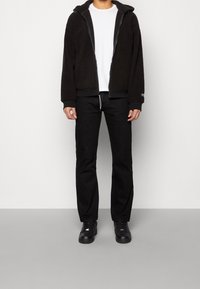 Person, der eine schwarze Fleecejacke mit Reißverschluss, ein weißes T-Shirt, schwarze Hosen und schwarze Turnschuhe trägt, steht auf einem grauen Boden vor einer weißen Wand.