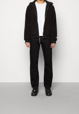 Personne portant une veste en polaire noire à zip, un t-shirt blanc, un pantalon noir et des baskets noires, se tenant sur un sol gris contre un mur blanc.