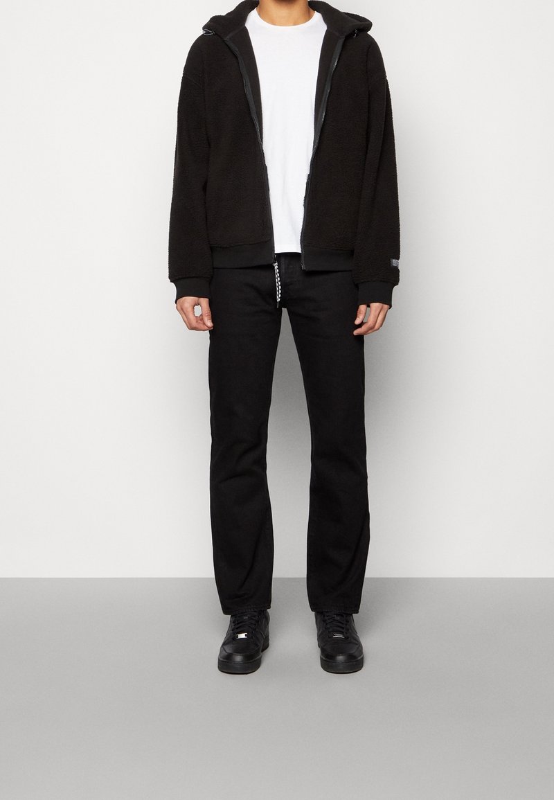 Person, der eine schwarze Fleecejacke mit Reißverschluss, ein weißes T-Shirt, schwarze Hosen und schwarze Turnschuhe trägt, steht auf einem grauen Boden vor einer weißen Wand.