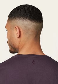 Man met kort, textuurhaar in een fade kapsel, gekleed in een donkerpaars shirt met een klein geborduurd logo op de bovenrug.