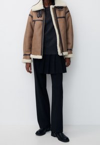 Giacca in shearling marrone con un rivestimento crema, dettagli neri, pantaloni a righe verticali neri e stivaletti neri alla caviglia. Design semplice e strutturato.