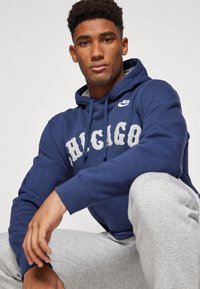 Mørkeblå hettegenser med "CHICAGO" i hvitt, med snøring i hetta og Nike-logo. Kombinert med lyse grå joggebukser.