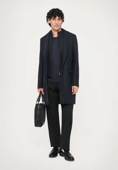 Joven con un abrigo oscuro, pantalones negros y zapatos negros, sosteniendo un maletín de cuero negro contra un fondo blanco liso.