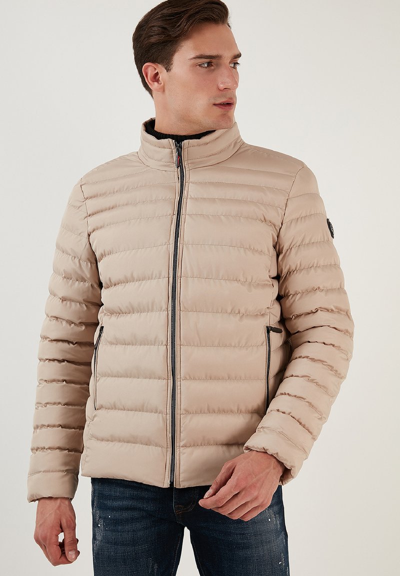 Beige pufferjakke med horisontalt quiltet design, høj krave, frontlynlås og sidelommer. Glat tekstur, letvægts og afslappet pasform.