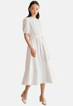 Vrouw draagt een witte midi-jurk met opgepofte korte mouwen, een ceintuur in de taille en beige schoenen met blokhak, staand op een effen achtergrond.