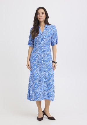 Nő kék-fehér mintás midi ruhában, rövid ujjakkal, világos, egyszínű háttér előtt áll, fekete magassarkú cipőt visel.